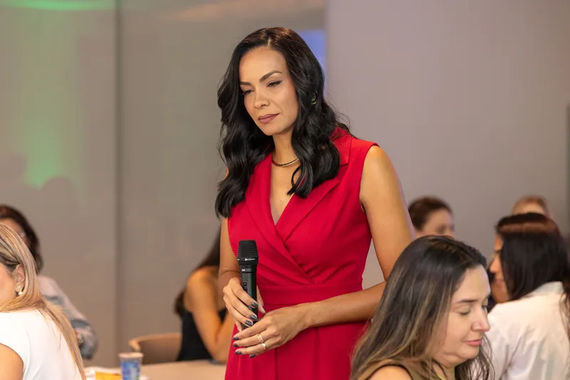 Daniela Rocha, de pé entre as mesas com o microfone nas mãos, observa a sala com expressão serena e firme.