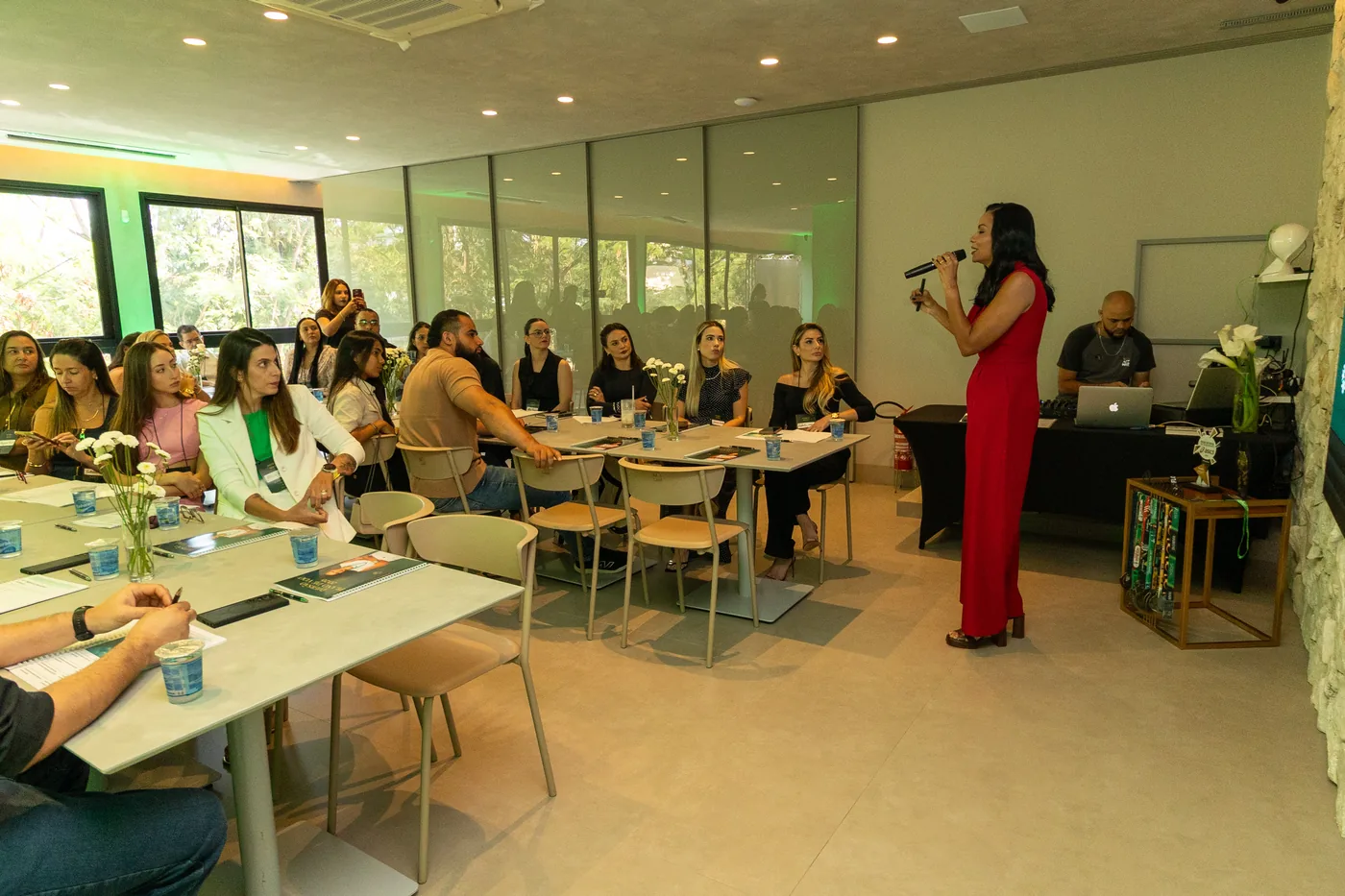 Daniela Rocha, de vestido vermelho e microfone na mão, conduz uma sala de participantes sentados em mesas.